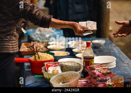 Nahaufnahme einer Frau, die ihre Hand mit vietnamesischem Bargeld ausstreckt, um eine Schüssel Nudeln für das Mittagessen in der Markthalle von Meo Vac zu kaufen. Stockfoto