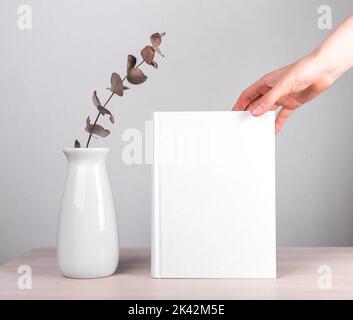 Abstraktes leeres leeres Buchcover, weiße Hardcover-Vorlage, weibliche Hand und Vase mit Eukalyptus auf Holztisch. Stockfoto