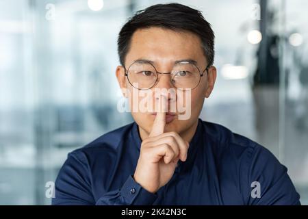 Mann in Brille und Casual Shirt Geschäftsmann Blick auf die Kamera arbeiten im modernen Bürogebäude asiatischen Investor hält Finger in der Nähe des Mundes anspruchsvolle Stille Stockfoto