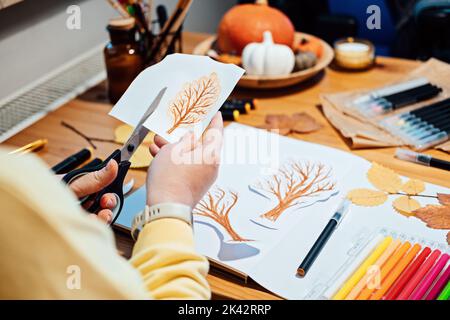 Herbstfertigkeit für Erwachsene. Gesichtsloses Porträt einer Frau, die mit Markern Herbstbäume zeichnet und Collage aus Papier ausschneidet. Stockfoto