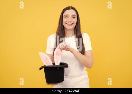 Hasenohren erscheinen aus dem Zauberhut. Porträt eines jungen Mädchens mit Hasenohren isoliert auf gelbem Hintergrund. Stockfoto