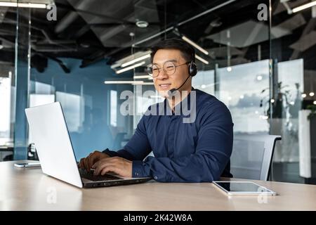 Glücklicher und lächelnder Mitarbeiter mit Headset für Videoanruf, asiatischer Techniker im Gespräch mit Online-Service-Benutzern, Geschäftsmann, der einen Laptop für den Online-Kundensupport verwendet. Stockfoto