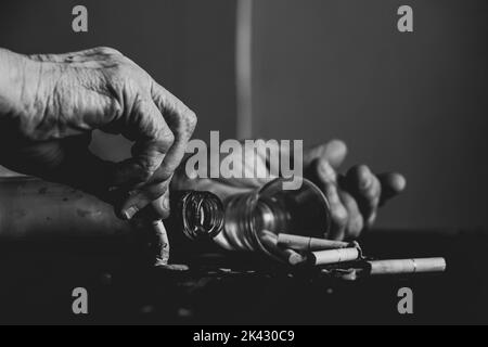 Zigaretten ein Glas und eine leere Flasche und die Hände einer alten Frau auf einem verbrannten schwarzen Tisch, weibliches Rauchen und Alkoholismus, schlechte Gewohnheiten Stockfoto