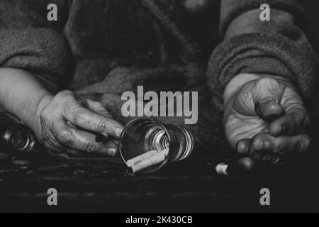 Zigaretten ein Glas und eine leere Flasche und die Hände einer alten Frau auf einem verbrannten schwarzen Tisch, weibliches Rauchen und Alkoholismus, schlechte Gewohnheiten Stockfoto