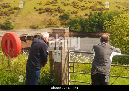 Zwei Männer ab 60 Jahren blicken über ein Tor und einen Zaun zum Meldon-Damm und See, Devon, England, mit einem Rettungsgürtel zur einen Seite an einem sonnigen Frühlingstag Stockfoto