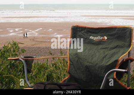 Nahaufnahme eines Campingstuhls mit Blick auf die Widemouth Bay, Cornwall, England, mit der Weite des Sands und zwei Spaziergängern und einem Hund Stockfoto