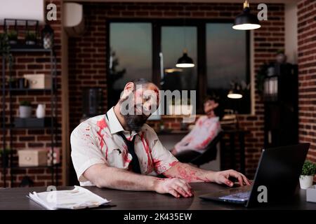 Porträt eines Zombie-Geschäftsmannes, der am Schreibtisch sitzt und im Firmenbüro gruselig und furchterregend aussieht. Gehirn essen grausames Monster mit blutigen Wunden arbeiten auf Laptop, schrecklich besessen Teufel. Stockfoto