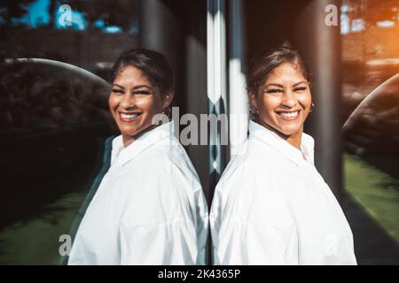 Ein Außenportrait einer fröhlichen reifen und großen hispanischen Frau mit einem schönen Lächeln, im weißen Hemd, lehnt sie sich an das Spiegelglas Stockfoto