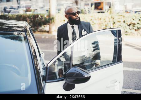 Ein Porträt eines zähen kahlen afrikanischen Geschäftsmannes mit einem schönen schwarzen Bart, in Sonnenbrille und einem taillierten modischen Kostüm, der zur Seite schaut, während er sich entblickt Stockfoto