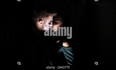 Halloween-Party, Nacht, erschreckender Mann in der Dämmerung, in den Strahlen des Lichts, mit einem schrecklichen Make-up in einem schwarzen Hexenkostüm. Dämon küsst das Mädchen. Der Werwolf küsst. Hochwertige Fotos Stockfoto