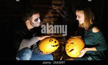 Datum im Stil von Halloween-Party, Nacht, Dämmerung, in den Strahlen des Lichts, Kerl mit einem Mädchen in Kostümen gekleidet und mit einem schrecklichen Make-up halten zwei große Halloween Kürbisse. Hochwertige Fotos