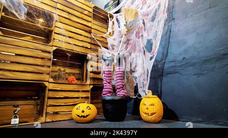 Halloween Party, Szenerie im Stil von Halloween mit Kürbissen, Spinnweben. In einem Kessel, einem Topf, ragen Frauenbeine in Stiefeln und einem Besen einer Hexe hervor. Hochwertige Fotos Stockfoto