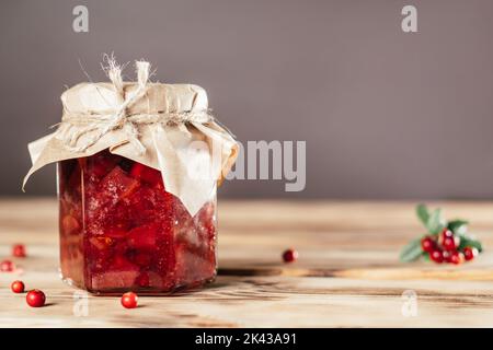 Glas mit hausgemachter Preiselbeeren- und Birnenmarmelade mit Bastelpapier auf Deckel auf Holzoberfläche neben frischen Preiselbeeren Stockfoto