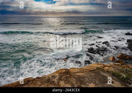 Sonnenstrahlen, die durch die Wolken eines bedrohlichen Sturms scheinen, während die Wellen des Pazifischen Ozeans über einem felsigen Vorgewende in Coolum, Sunshine Coast, Qu, brechen Stockfoto