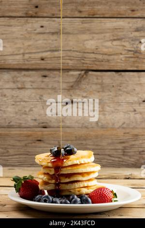 Vertikales Bild von Ahornsirup, der auf Pfannkuchen und Heidelbeeren auf Holz mit Kopierfläche gießt Stockfoto