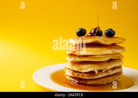 Horizontales Bild von Ahornsirup, der auf Pfannkuchen und Heidelbeeren gießt, auf Gelb mit Kopierfläche Stockfoto