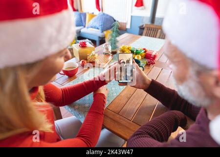 Kaukasisches Paar in santa Hüte macht Smartphone weihnachten Videoanruf mit verschiedenen älteren Freunden. weihnachten, Festlichkeit und Kommunikationstechnik. Stockfoto