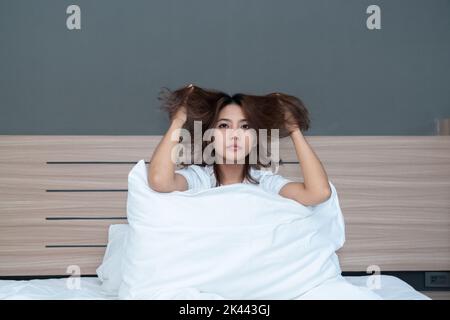 Junge asiatische Frauen schläfige junge Frau mit zerzausten Haaren sitzt auf dem Bett zu Hause Stockfoto