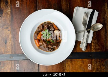 Eine weiße Schale mit Boeuf Buorguignon - französischem Rindereintopf - auf einem rustikalen Holztisch, mit Serviette und Besteck Stockfoto