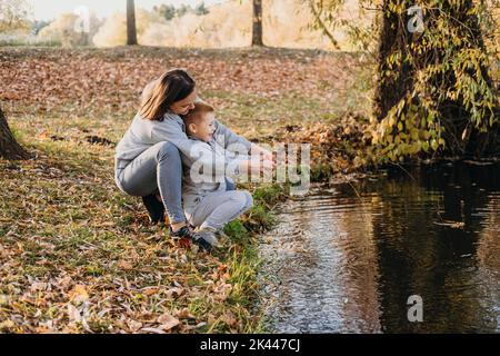 Mutter mit Kindern fischen mit einem Stock am Rande eines Sees im Herbstpark. Wilde Natur und Wasserspaß im Herbsturlaub. Camping und Angeln Stockfoto