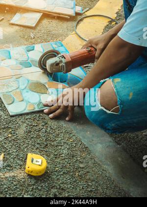 Hände von Bauarbeitern, die einen Winkelschleifer zum Schneiden der Fliesen auf der Baustelle verwenden. Ein Arbeiter schneidet beige Fliesen mit einer Kreissäge. Schnitt Stockfoto