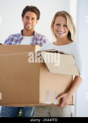 Umzug in ein eigenes Haus. Porträt einer lächelnden Frau, die einen braunen Karton trägt, während ihr Freund im Hintergrund einen Karton trägt. Stockfoto