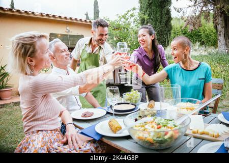 Fröhliche Erwachsene Freunde lächeln und Weingläser bei der Feier im Hinterhof des Hauses toasten. Zwei reife Männer und drei Frauen haben am Wochenende Spaß miteinander Stockfoto