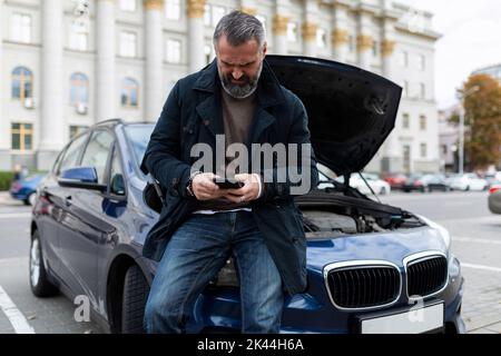 Ein erwachsener Mann ruft neben einem kaputten Auto mit offener Motorhaube den technischen Support für die Autoreparatur an Stockfoto