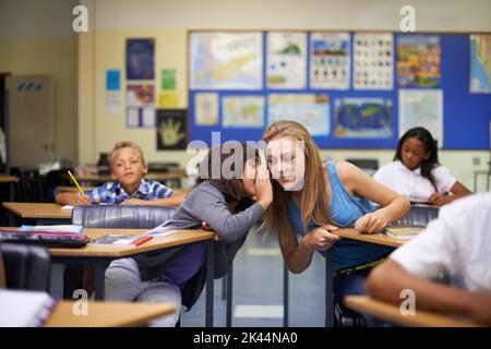 Haben Sie schon gehört. Ein Teenager-Mädchen flüstert ihren Freunden mitten in einem Klassenzimmer ins Ohr. Stockfoto