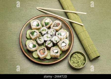 Sushi und Gurken in Scheiben auf Keramikplatte. Grüne Bambusmatte für Sushi, Essstäbchen und Wasabi in einer Schüssel. Speicherplatz kopieren. Flach liegend. Grüner Hintergrund. Stockfoto
