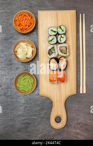 Auswahl an Sushi auf Schneidebrett. Eingelegter Ingwer und Karotten, Wasabi in Holzschüsseln. Essenssticks. Flach liegend. Brauner Hintergrund. Stockfoto