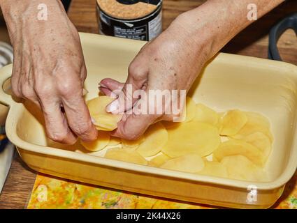Schritt-für-Schritt-Rezept für Kartoffelgratin. Die Hände stapeln Kartoffelscheiben in eine Glasbackform. Kartoffelauflauf Stockfoto