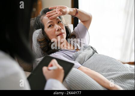 Eine ältere asiatische Patientin befindet sich im Aufwachraum und wird von ihrem Arzt untersucht. Eine Ärztin spricht, überprüft und zeichnet den Strom auf Stockfoto