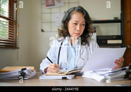 Professionelle asiatische Ärztin im Alter, die sich beim Lesen einer medizinischen Forschungsarbeit oder Schreiben eines medizinischen Chirurgie-Fallergebnisses auf einer medizinischen Arbeit in h Stockfoto