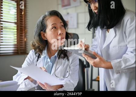 Professionelle leitende asiatische Ärztin zeigt und erklärt ihrem Assistenten vorab medizinische Informationen über die Forschung. Stockfoto