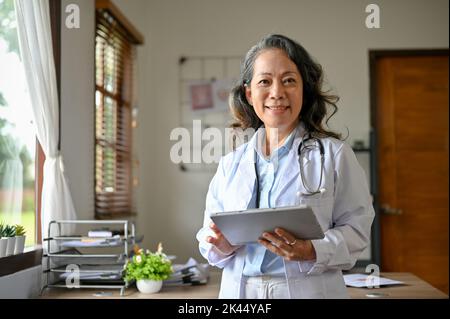 Die professionelle und erfolgreiche Senior Asian Ärztin in weißem Kleid hält ein digitales Tablet und steht in ihrem Büro. Stockfoto