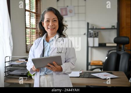 In ihrem Klinikbüro steht eine professionelle und selbstbewusste weibliche leitende asiatische Ärztin in weißem Kleid mit Stethoskop und digitalem Tablet. Stockfoto