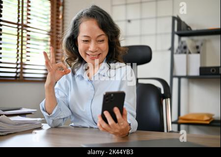 Die schöne und glücklich gealterte asiatische Geschäftsfrau sitzt an ihrem Schreibtisch und führt mit ihrem Kollegen einen Videoanruf über ihr Smartphone, wodurch sie Okay Hand si macht Stockfoto
