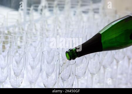 Nahaufnahme einer Champagnerflasche, die Champagner in eine Sektflöte vor einem verschwommenen Glas im Hintergrund pochelt Stockfoto