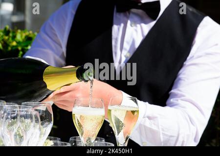 Nahaufnahme einer Champagnerflasche, die Champagner auf einer Sektflöte gegen einen verschwommenen Kellner auf einer Party umschellt Stockfoto