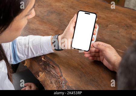 Modernes vertikales Smartphone-Modell, über der Schulteransicht des modernen vertikalen Smartphone-Modells. Das Paar sitzt auf einem Holztisch und sieht aus wie ein leerer Bildschirm. Stockfoto