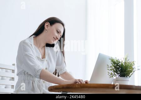 Lächelnde Frau, die an einem Laptop arbeitet und auf ihrem Smartphone spricht. Stockfoto