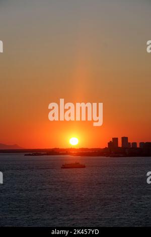 Die helle Sonne im roten Sonnenuntergang über dem Ägäischen Meer in Izmir, Türkei mit Gebäuden im Hintergrund Stockfoto