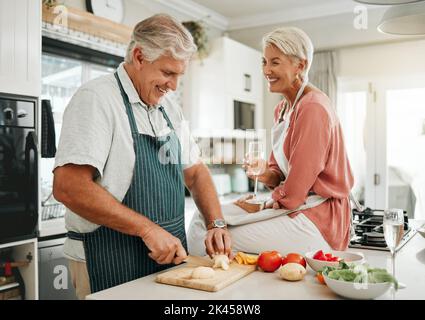 Ein glückliches Seniorenpaar, das in der Küche gesundes Essen kocht und Champagner trinkt, während sie den Ruhestand genießen. Ältere Frau, die auf der Theke sitzt, Mann Stockfoto