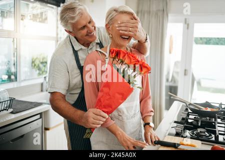 Das ältere Paar, das Augen und Blumen bedeckt, überrascht, als der Mann seiner Frau an einem Jahrestag, Geburtstag oder Valentinstag einen Rosenstrauß in der Küche schenkte. Glücklich Stockfoto