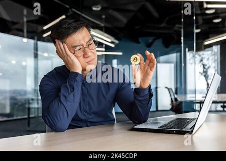 Frustrierter asiatischer Geschäftsmann, Investor, der Krypto-Währung-Bitcoin in den Händen hält, Mann, der im modernen Bürogebäude arbeitet, scheitert an finanziellen Investitionen. Stockfoto