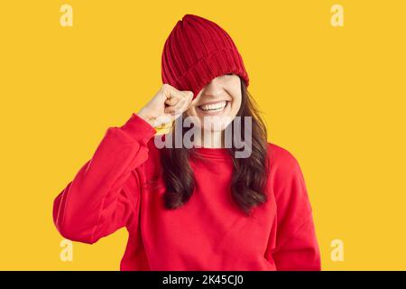 Lustige nette junge Frau lacht und versteckt ihre Augen, indem sie einen gestrickten Hut über sie zieht. Stockfoto