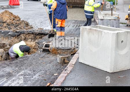 Eine Gruppe von Straßenarbeitern repariert an einem Herbsttag auf einer Straße die Kanalisationslüfte und wechselt alte Betonschächte gegen neue. Stockfoto