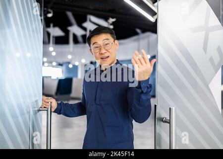 Der Chef des asiatischen Geschäftsmannes lädt zu seinem Team ein, der Mann schaut auf Kamera und Wellen, der Manager in Brille und Hemd arbeitet im modernen Bürogebäude. Stockfoto