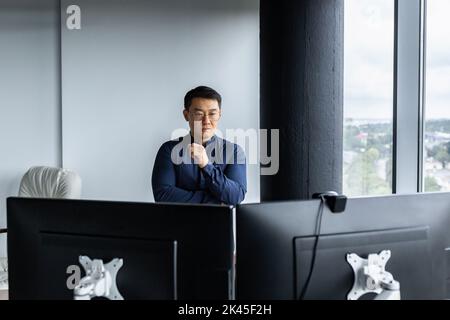 Ein seriöser und denkender asiatischer Ingenieur, der zwei Monitore ansah, ein Nahaufnahme-Foto von einem Büromitarbeiter, einem Mann, der im modernen Bürogebäude arbeitet, einem Geschäftsmann, der eine Brille und ein legeres Hemd trägt. Stockfoto
